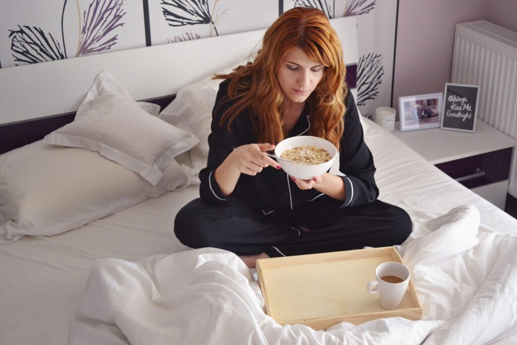 A young woman sitting on a bed with an eiderdown comforter while eating a bowl of cereal